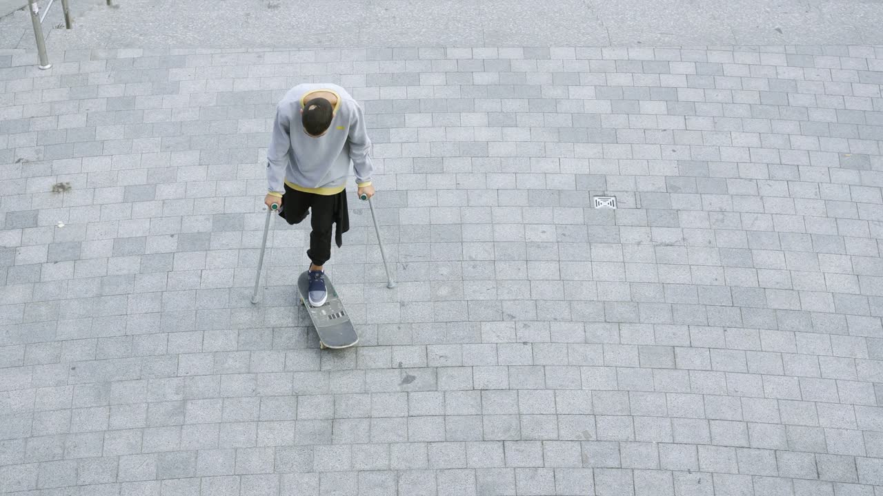 Ovenfra: Mann med ett ben på krykker spinner på skateboardet sitt og gjør triks.