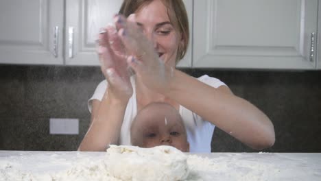 Young-mother-and-her-little-child-preparing-dough-on-the-table,-clapping-hands-and-enjoying-their-time-together.-Family-girls