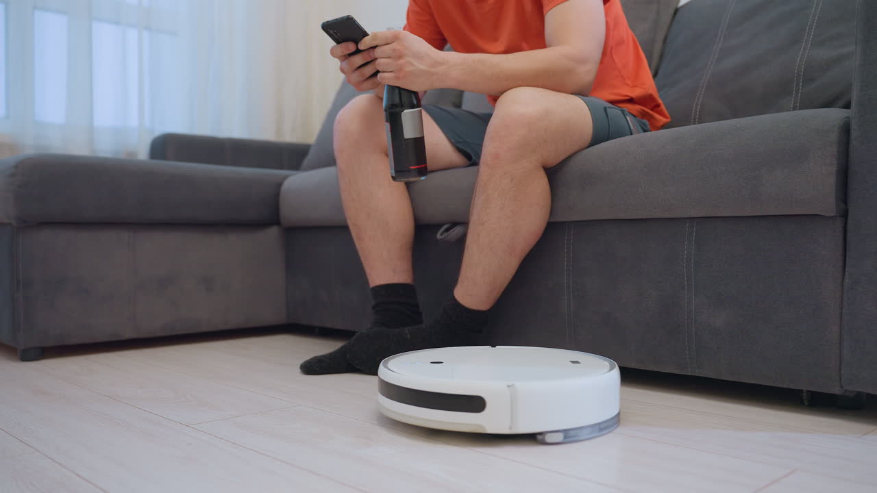 Man sitting on couch operates phone as robot vacuum delivers bottle of drink, he collects it while device turns and moves away, showcasing convenience and creative use of smart home technology
