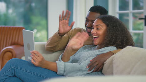 Pareja-Joven-Relajándose-En-El-Sofá-De-Casa-Usando-Una-Tableta-Digital-Para-Hacer-Videollamadas-Juntos