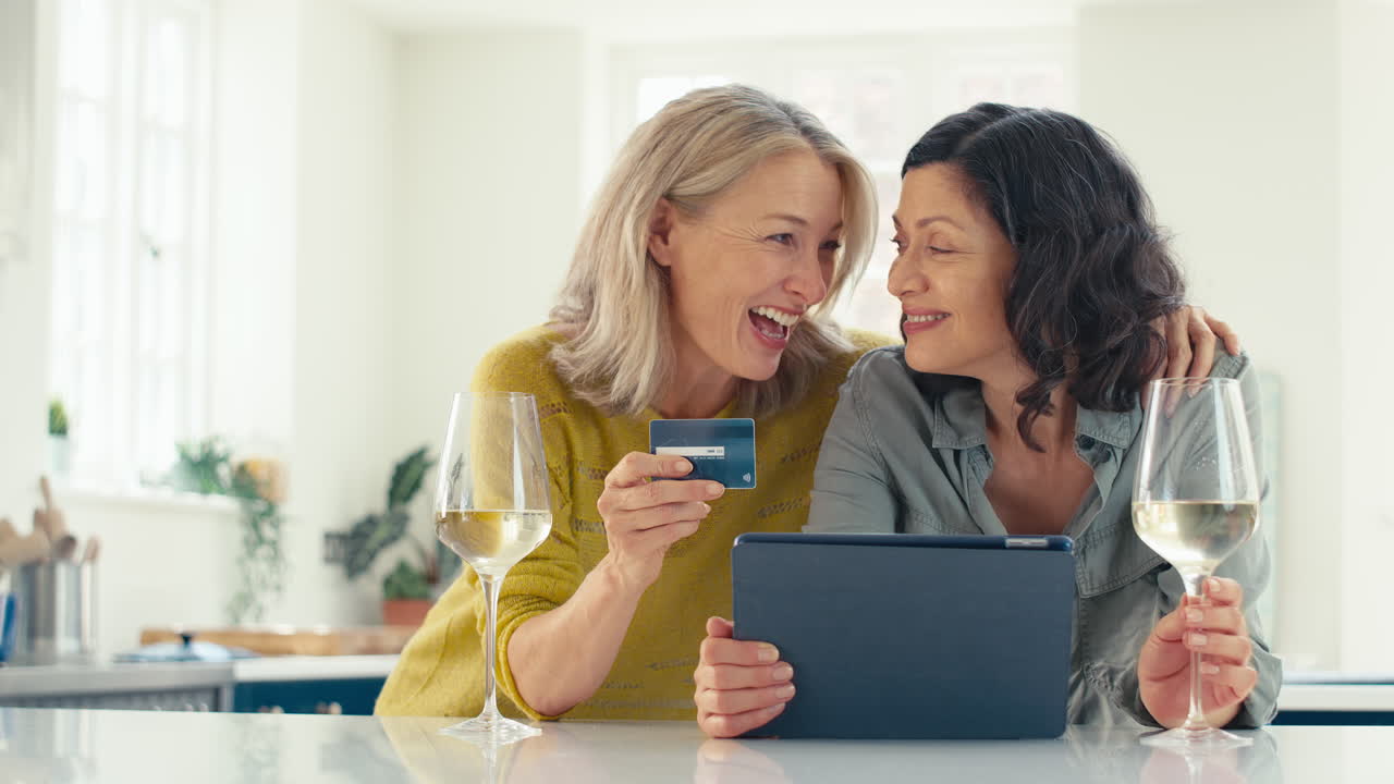 Same Sex Mature Female Couple With Credit Card Using Digital Tablet At Home To Book Holiday Or Shop