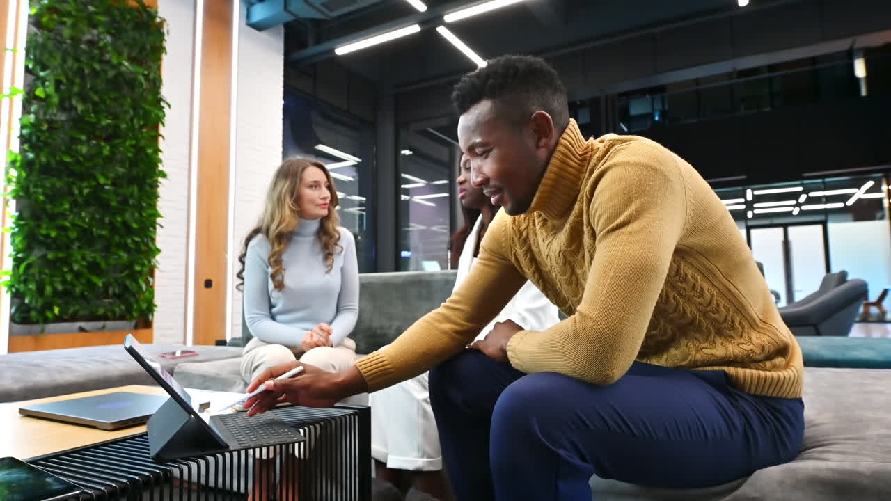 Caucasian and black female workers discussing business while a black man is using a tablet, sitting on a sofa in an office. Slow motion