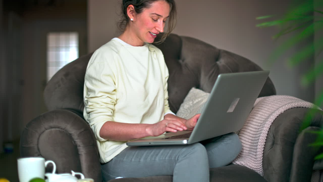 Happy caucasian woman sitting on a chair and working on her laptop. Table with tea and fruits nearby