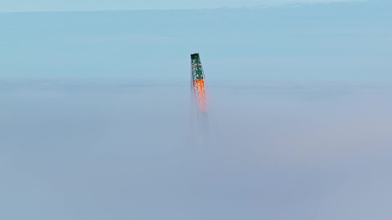 The top section of a tall construction crane rises above a dense layer of clouds, striking aerial view - machinery float in a soft, misty atmosphere