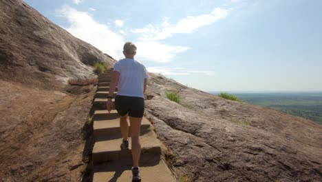 hyper-lapse tracking from behind a blonde western tourist as she climbs up stairs in the side of a large granite mountain in east africa with incredible landscape views