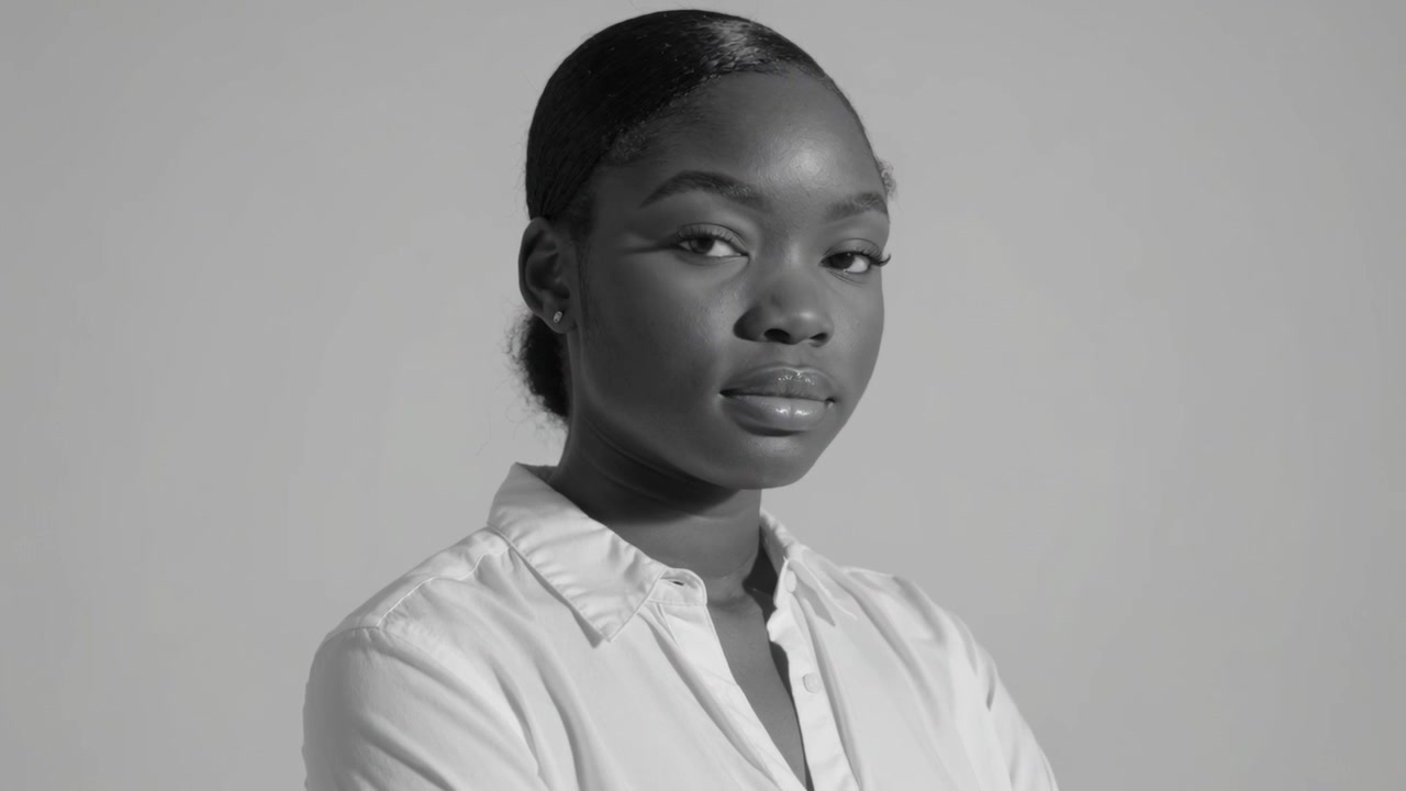 La cámara comienza con una modelo posando en un estudio, cambiando la mirada y sonriendo, con camisa blanca abotonada y tachuelas.