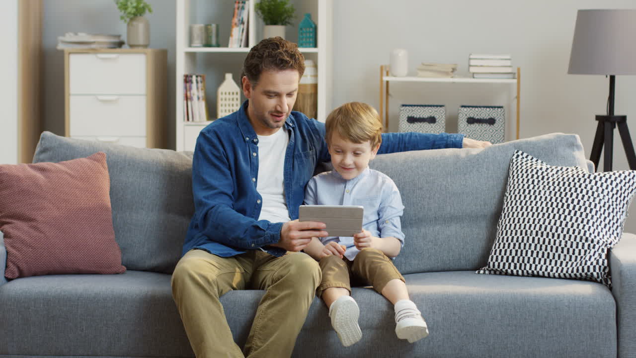 Handsome Father Sitting On The Gray Couch And Hugging His Cute Little Son While They Watching Something On The Tablet Computer In The Living Room