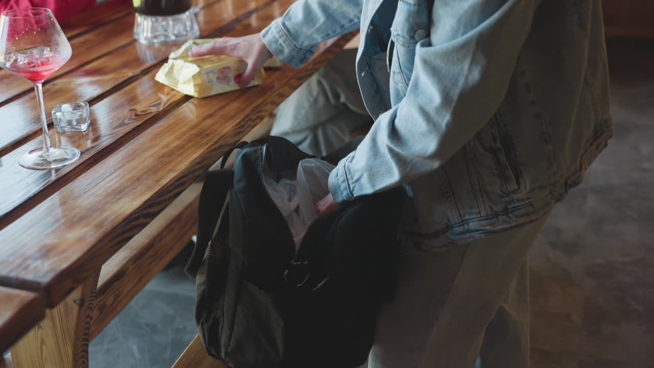 Close up of lady in denim jacket unpacking biscuit packs from black bag and placing them on polished wooden table next to half-full wine glass and candles, during casual indoor gathering in daylight