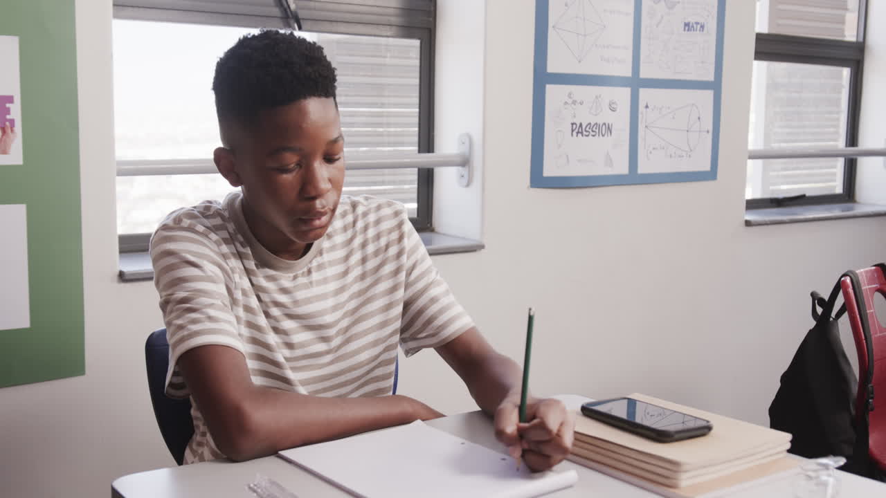 In school, African American teenage boy writing in notebook at desk, focusing on his work