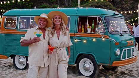 happy mature couple holding cocktails while standing next to a vintage van arranged as a bar on a beach, enjoying a summer evening together