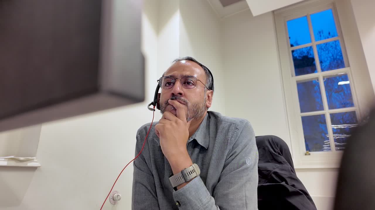 Uomo dell'Asia meridionale con cuffie che lavora alla scrivania in ufficio con laptop e monitor, immerso nei suoi pensieri mentre riflette sulla strategia aziendale e sulla risoluzione dei problemi nell'area di lavoro