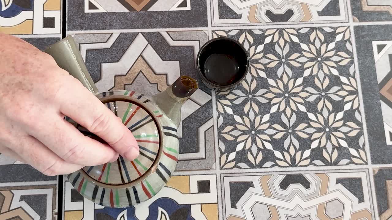 En hånd åbner låget på en stribet keramisk tekande for at afsløre teblade på mosaikfliser.