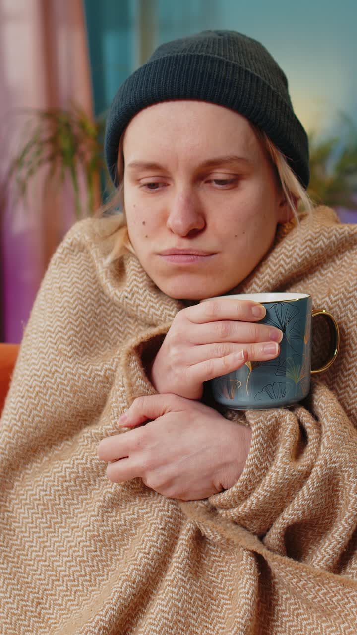 Ill sick unhealthy woman freezes from the cold feels bad covered by warm blanket drinks tea at home