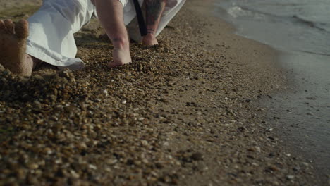 Nahaufnahme-Der-Beine-Eines-Mannes,-Der-Sich-Auf-Nassem-Sand-Ausdehnt.-Kämpferhocke-Beim-Training-In-Der-Natur.