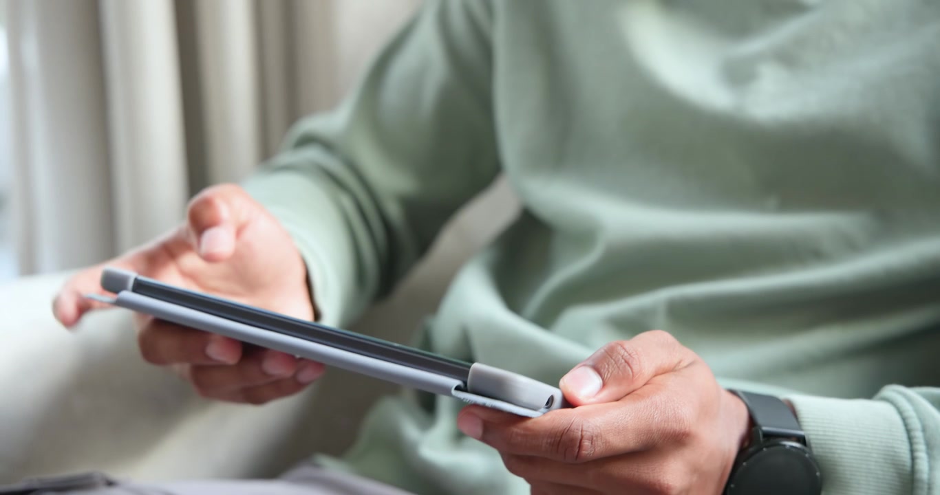 Adult African American man preparing to stow tablet, holding, folding grey folio on couch by window