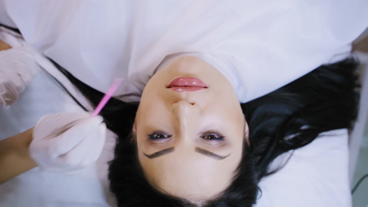A beautiful woman with black hair is lying on a couch on the procedure of permanent makeup lips. Master disinfects the area of pigment application. Close-up.