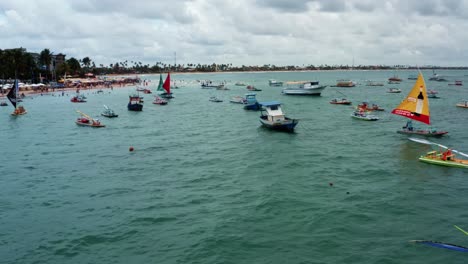 Dolly-in-aerial-drone-shot-of-the-Porto-de-Galinhas-or-Chicken-Port-beach-with-anchored-sailboats-and-tourists-swimming-in-the-crystal-clear-ocean-water-in-Pernambuco,-Brazil