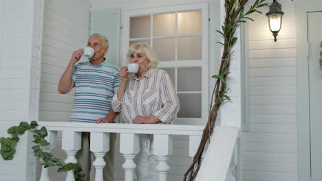 Senior-elderly-Caucasian-couple-drinking-coffee,-embracing-in-porch-at-home.-Mature-happy-family