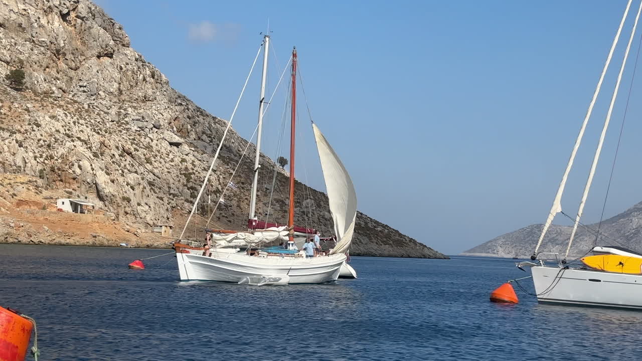 Greece,Kalimnos Island,Palionnisos, a white classical sailboat is leaving the bay, passing in between other sailboats in vaults and people on the boat is setting its front sail while leaving the bay.