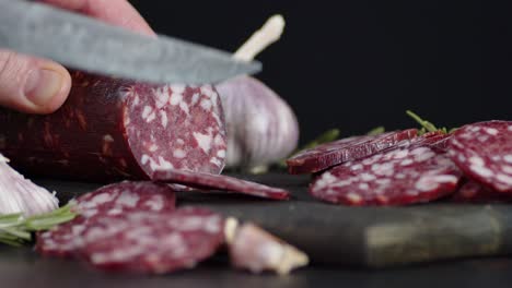 male hand cut a stick of salami on a cutting board.