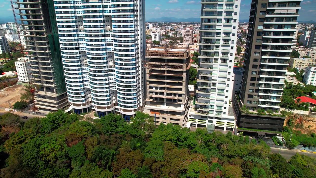 Ripresa aerea con drone dall'alto che avanza e si sposta a destra, parallela a grattacieli residenziali di lusso e costruzioni edilizie lungo un viale alberato