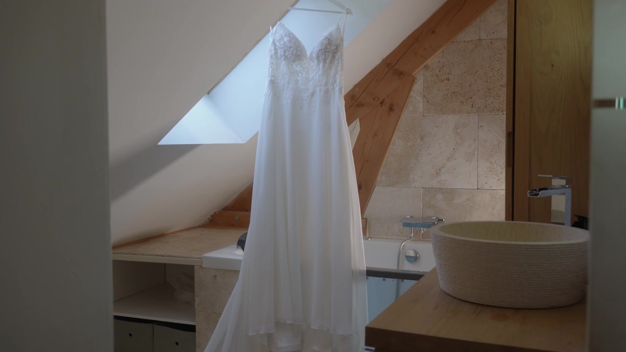 Slow push in revealing a white lace wedding dress hanging under a roof windows in a rustic attic bathroom with skylights and wooden beams. soft diffused light.