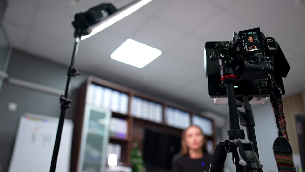 Modern professional camera on tripod is filming a woman speaking in the office. Low angle view at the camera and daylight lamp. Blurred backdrop.
