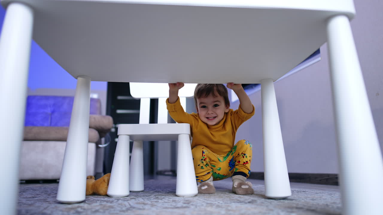 Cute smiling kid squatting near the desk. Playful cheerful baby boy in his room.