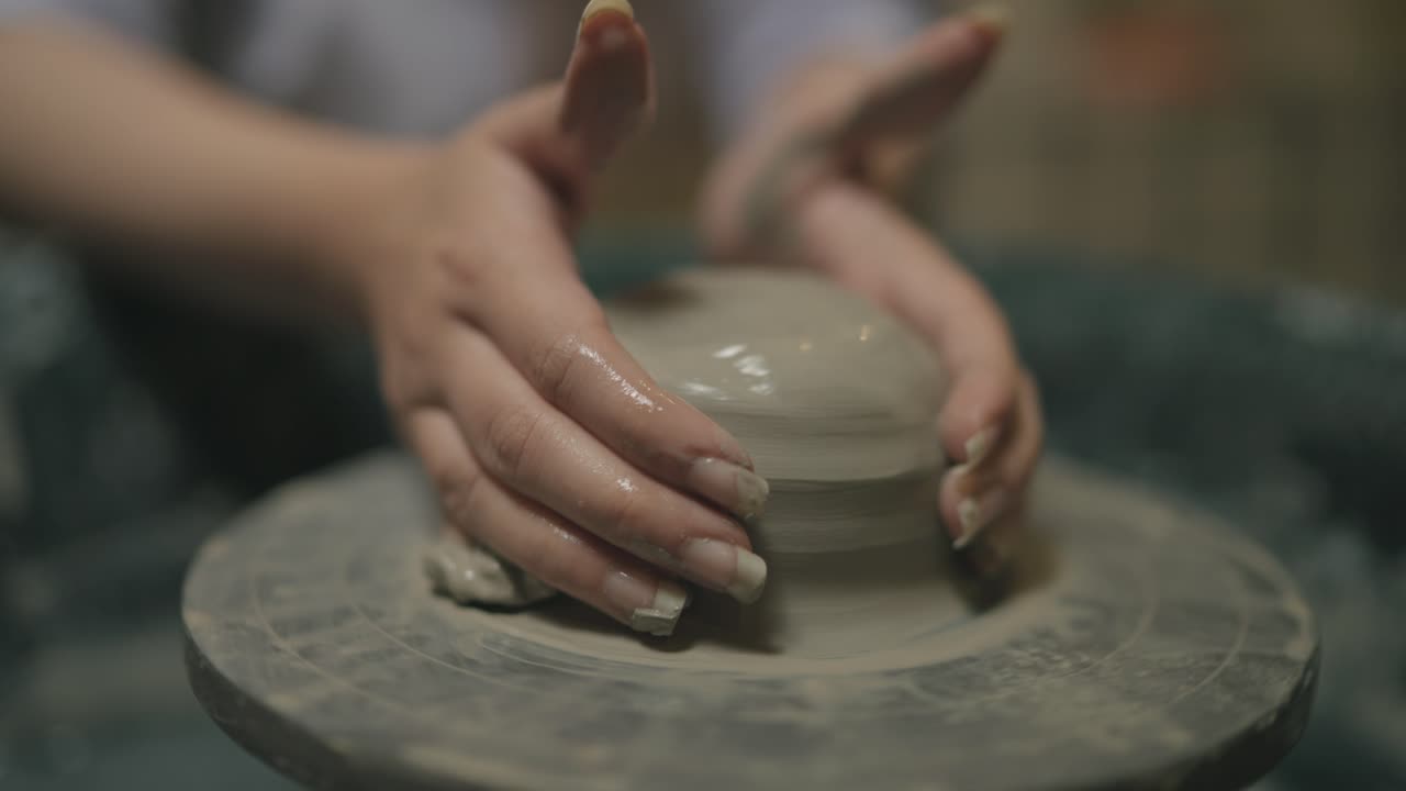 Close-up captures artisan's hands refining clay cylinder on pottery wheel, fingertips smoothing wet surface as the form takes shape under soft studio light, showing precision of ceramic crafting