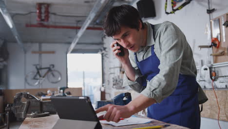 propietario de un negocio masculino en un taller usando una tableta digital y haciendo llamadas por teléfono móvil