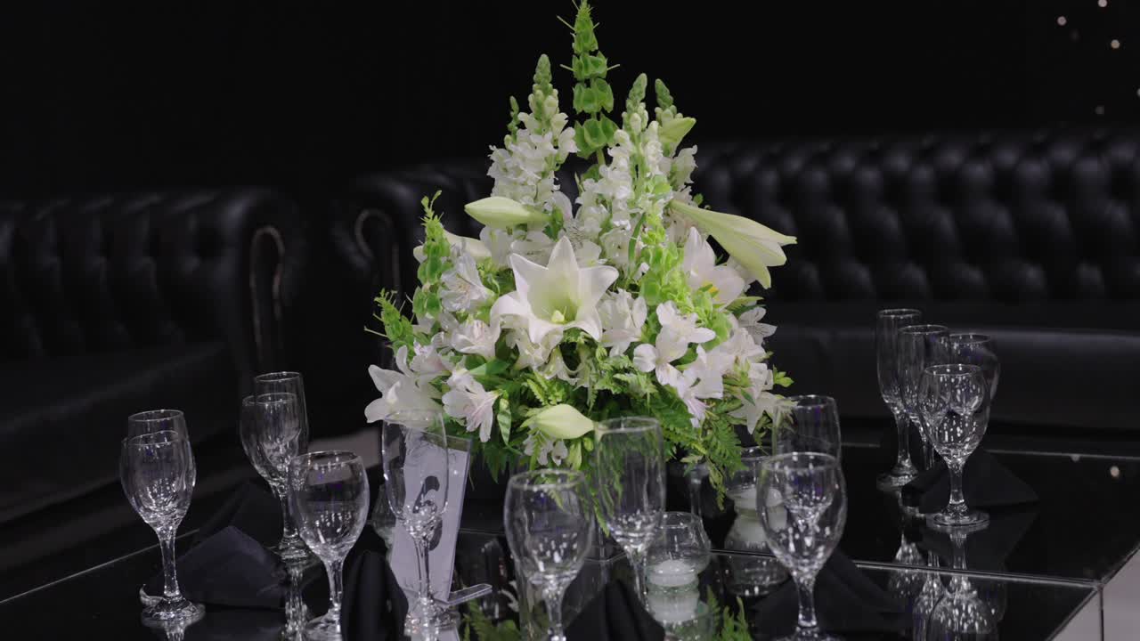 Bryllupsborddekking med hvitt blomsterarrangement i midten, med Lilium candidum og dekorativt grønt, omgitt av krystallglass på en reflekterende overflate i et elegant innendørs lokale, med kamera som zoomer inn og roterer.