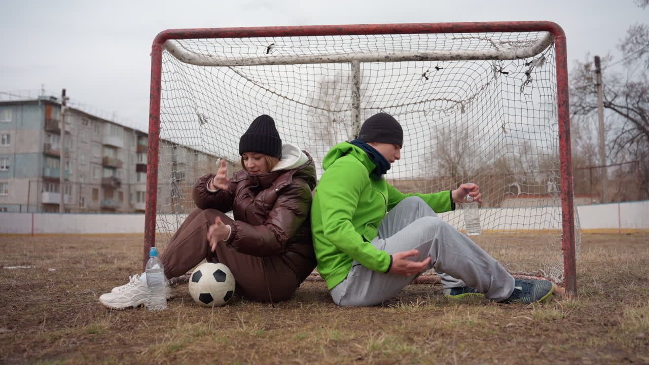 진흙투성이 잔디 위에 놓인 축구공과 낡은 그물, 축구 골대 안에 앉아 있는 두 명의 백인 십 대가 있어요. 한 명은 앞으로 손을 뻗고 다른 한 명은 쉬고 있으며, 비니 모자와 패딩 재킷을 입고 있어요. 황량한 도시 외곽, 흐린 하늘입니다.