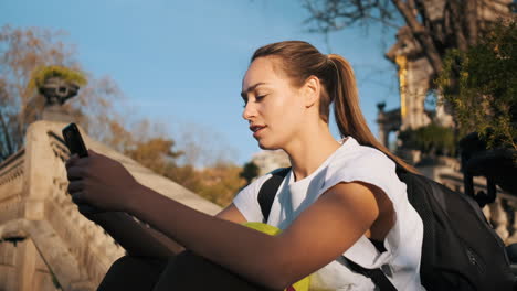 Mujer-Con-Fútbol-Y-Mochila-Felizmente-Usando-Un-Teléfono-Inteligente-Durante-El-Descanso-En-El-Parque-De-La-Ciudad.