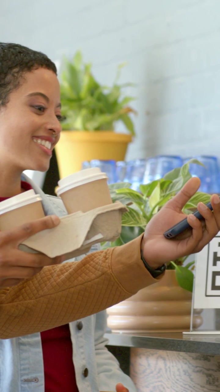 Video vertical: Mujer sonriente con ropa vaquera sosteniendo tazas en una cafetería, hombre acercando el teléfono para escanear un código QR y pagar.