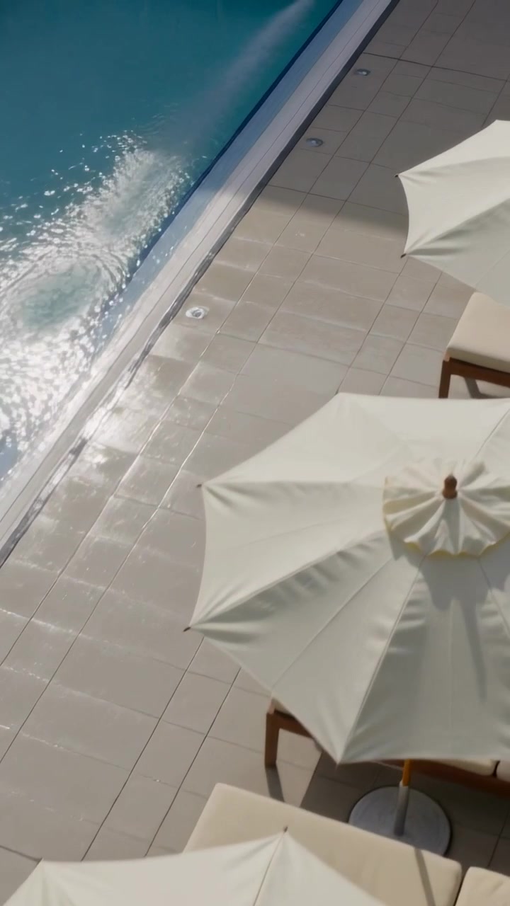 Vertical video: Traveling wavelets splashing pool edge, casting parasol shadow on tiled resort deck