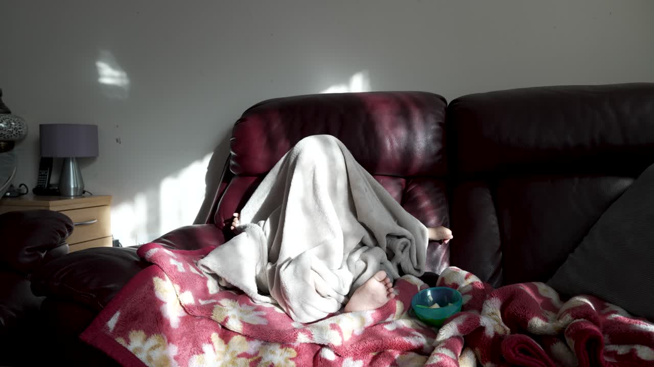 Funny British Asian Toddler Pulling Blanket Over Himself Sitting On Sofa In Living Room With Morning Sunlight Coming Through Window