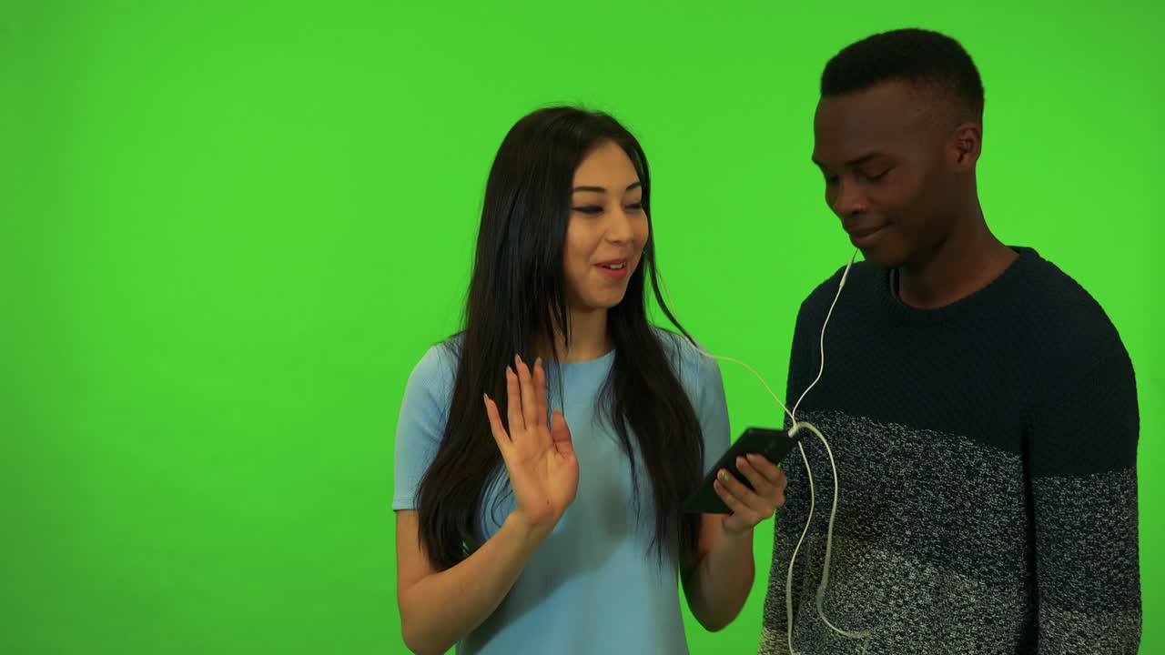 una joven asiática y un joven negro escuchan música en un teléfono inteligente - estudio de pantalla verde