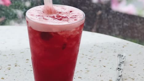 a vibrant red drink with a straw sits on a speckled outdoor table, surrounded by a blurred floral background.