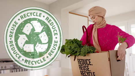 biracial woman in hijab carrying box with vegetables in kitchen over recycling sign