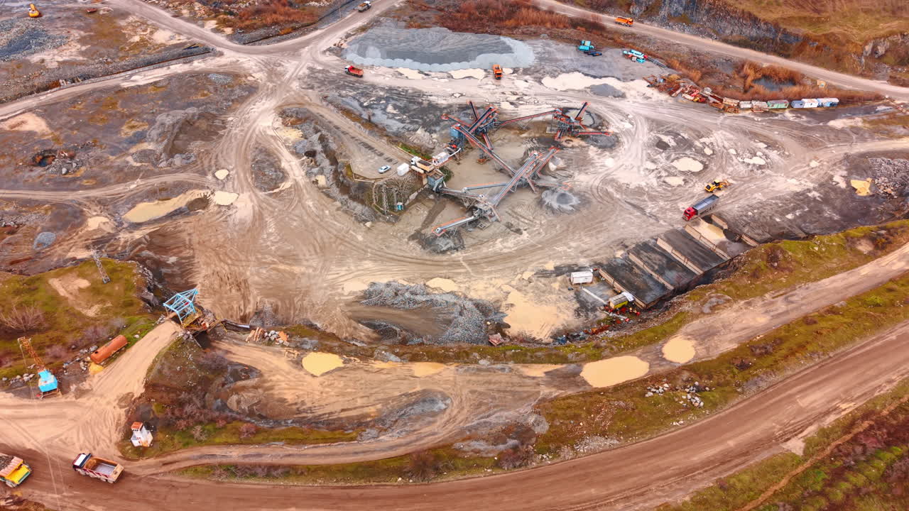 Industrial machinery for stone crushing and sorting in open pit. Wide aerial view of a granite quarry processing area with conveyors, trucks, and muddy ground