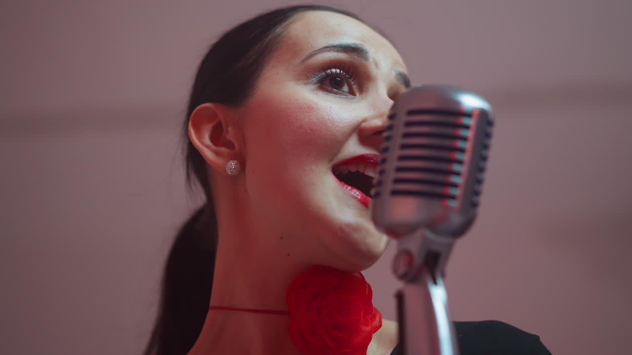 은 입술과 목에 리본을 은 기쁜 여성 보컬이 빈티지 마이크에 열정적으로 노래하며 그녀의 표현력과 활기찬 에너지를 포착합니다.