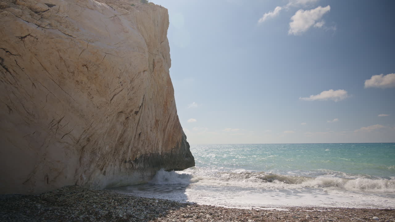 Dramatic view of a large cliff face meeting the sea at Aphrodite's Rock in Cyprus. Waves gently hit the shore under bright sunlight and blue sky