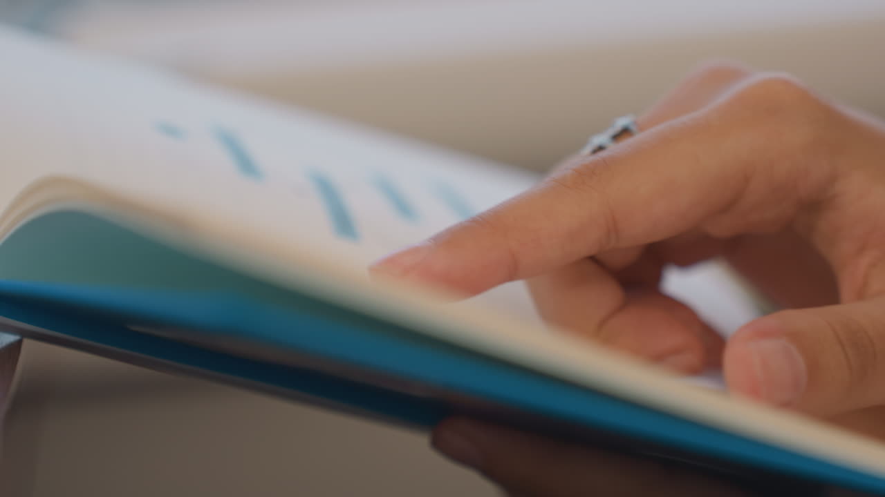 Closeup hands flipping blue notebook pages during focused planning session analyst reviews flowchart and charts, traces diagram with fingertip, turns cover, notices subtle ring on finger, natural