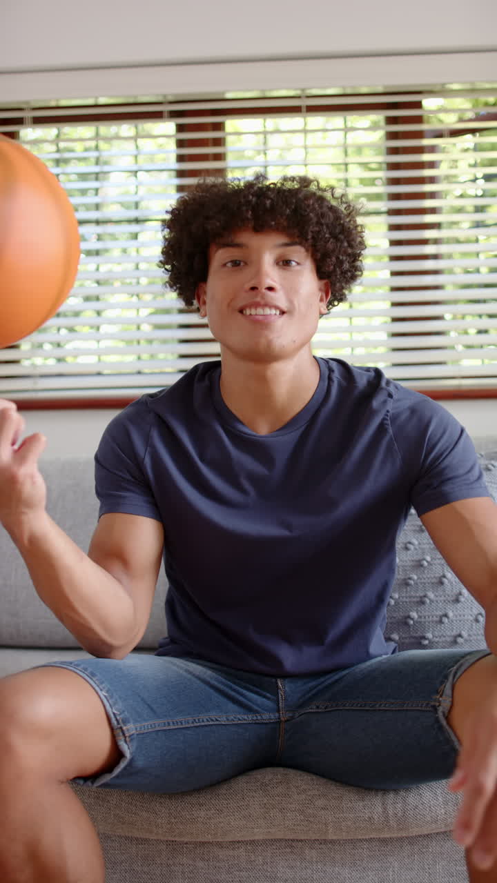 Video verticale: Giovane uomo che fa girare una palla da basket sul dito mentre è seduto sul divano a casa.