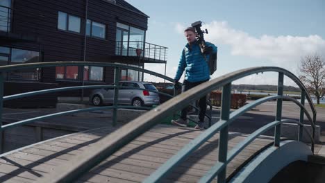 medium wiide shot van camera man in blauwe jas wandelen met cinema camera op een statief terwijl op zoek naar goede composities voor de volgende scène in een kleine stad