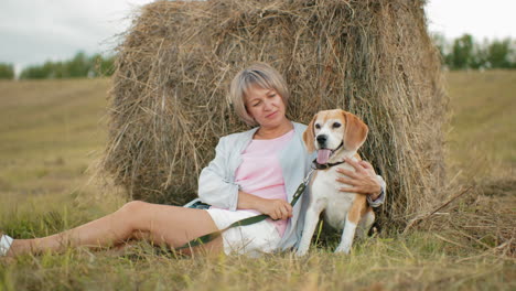 middle-aged woman wearing sunglasses sits against hay bale, rubbing dog back as it pants happily, rolling hills, golden grass, and cloudy sky