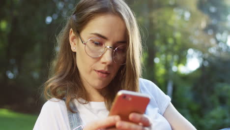 close-up view of a young caucasian woman in glasses lying on a blanket in the park tapping and scrolling on a smartphone