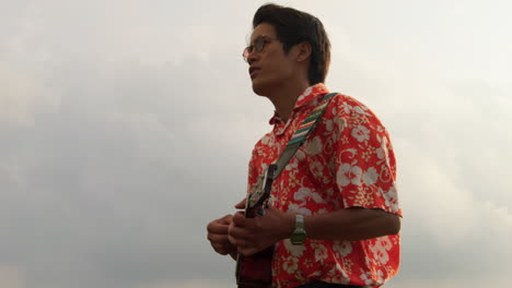 close up shot of an asian man standing on top on a mountain singing a song while playing ukulele on a cloudy day