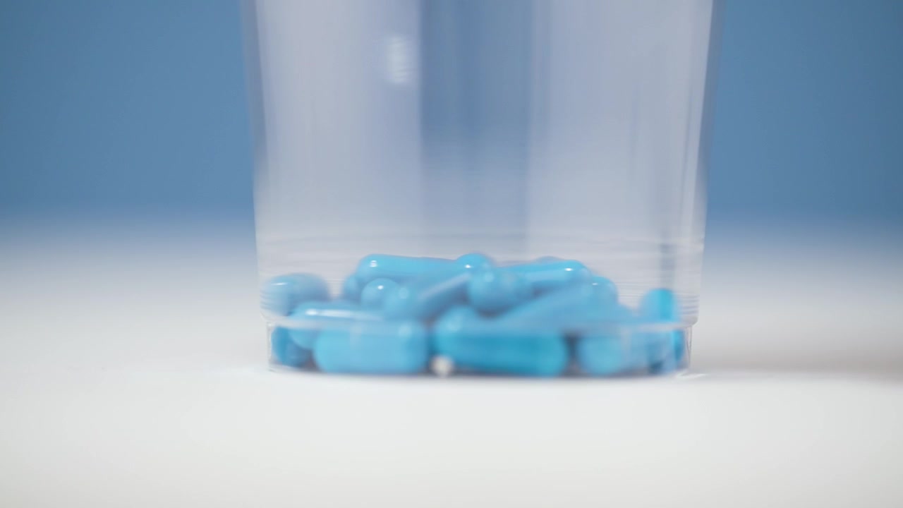 Una scatola trasparente piena di pillole blu viene delicatamente posizionata su una superficie bianca e pulita in slow motion 4K. Lo sfondo blu sereno esalta i toni vivaci e la finitura lucida delle pillole e della scatola