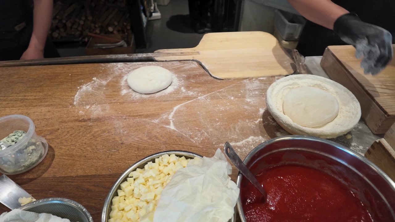 Ripresa dall'alto di uno chef con guanti neri che lavora l'impasto della pizza tra ingredienti freschi e farina